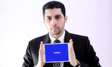 Disgruntled-looking man holds iPad with Facebook logo showing on its screen.