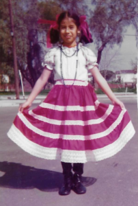 Little Mexican American girl wearing traditional norteño costume: white blouse, red and white skirt, red ribbons tied around braids.